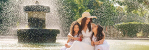 A woman with two children sitting by a fountain, enjoying a sunny day, surrounded by lush greenery and the soothing sound of water.
