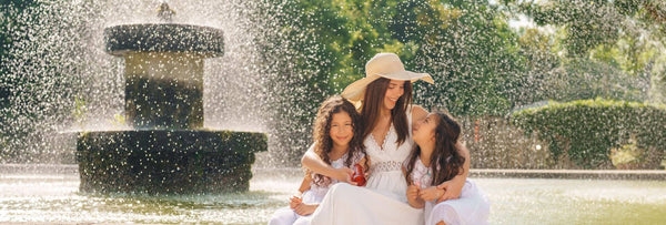 A woman with two children sitting by a fountain, enjoying a sunny day, surrounded by lush greenery and the soothing sound of water.