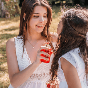 Elegant woman in a white lace dress sharing an exquisite fragrance experience with a young girl in a sunlit garden, evoking Maison Maïssa's charm.