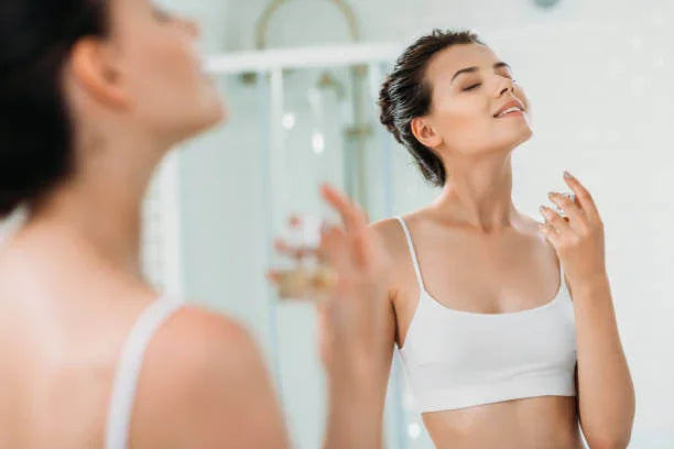 Young woman in white crop top applying perfume in bright bathroom mirror