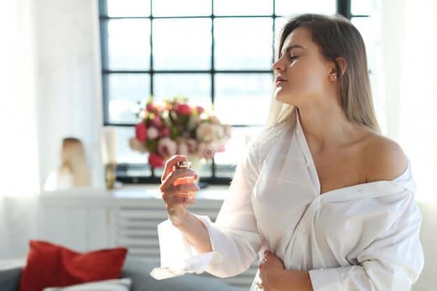 Woman in white shirt applying perfume indoors near window with floral bouquet
