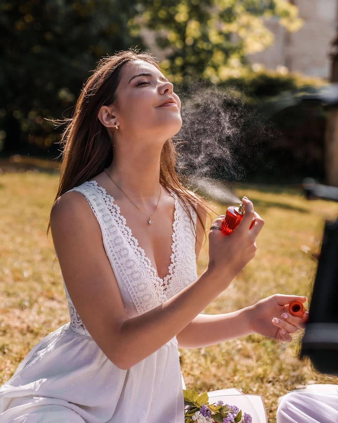 Young woman in white lace dress spraying perfume outdoors on sunny day with flowers nearby