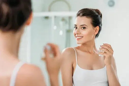 Smiling young woman in white tank top holding a glass of water, looking at her reflection in a bathroom mirror