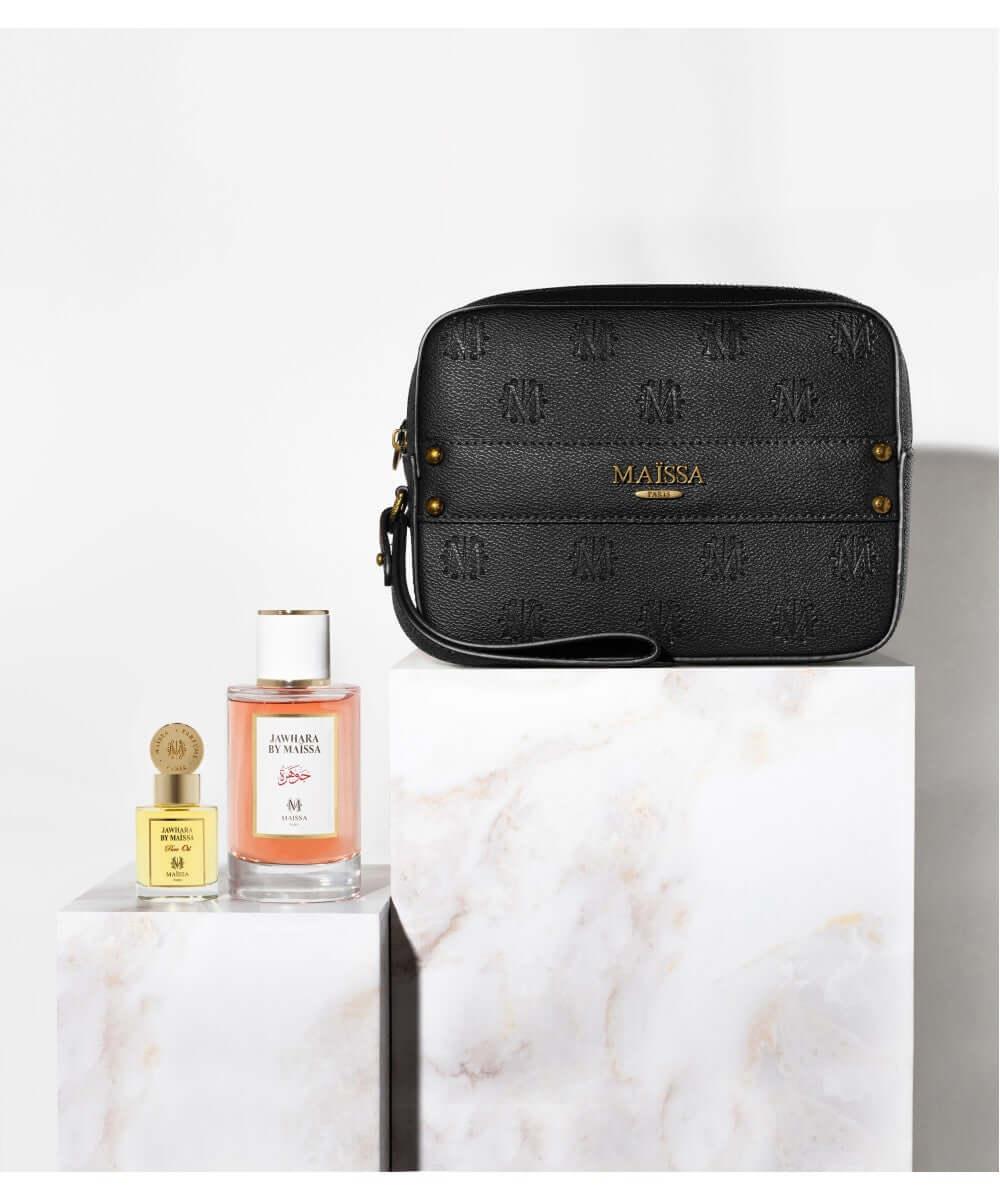 Black Maissa embossed leather clutch bag with gold logo, displayed with Jawhara by Maissa perfumes on marble pedestals