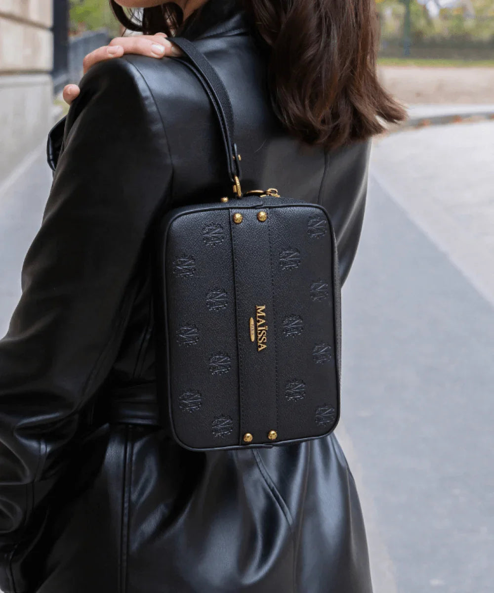 Black Maissa leather bag with embossed logo and gold details, alongside two Destin des Anges perfume bottles on white marble pedestals